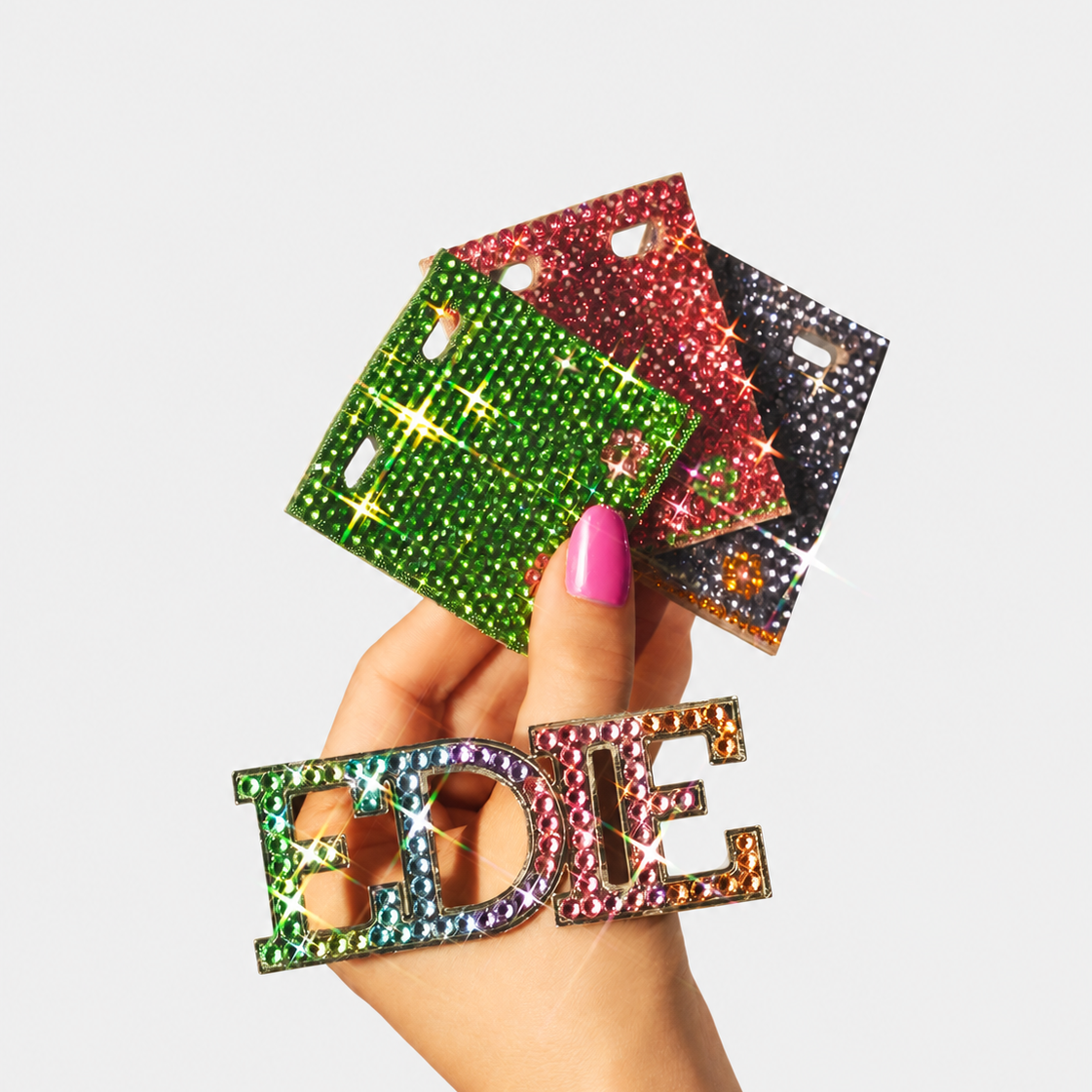 A hand with pink nail polish holds three square rhinestone-covered letter cards and a matching rhinestone letter 'E' in front of a light gray background.