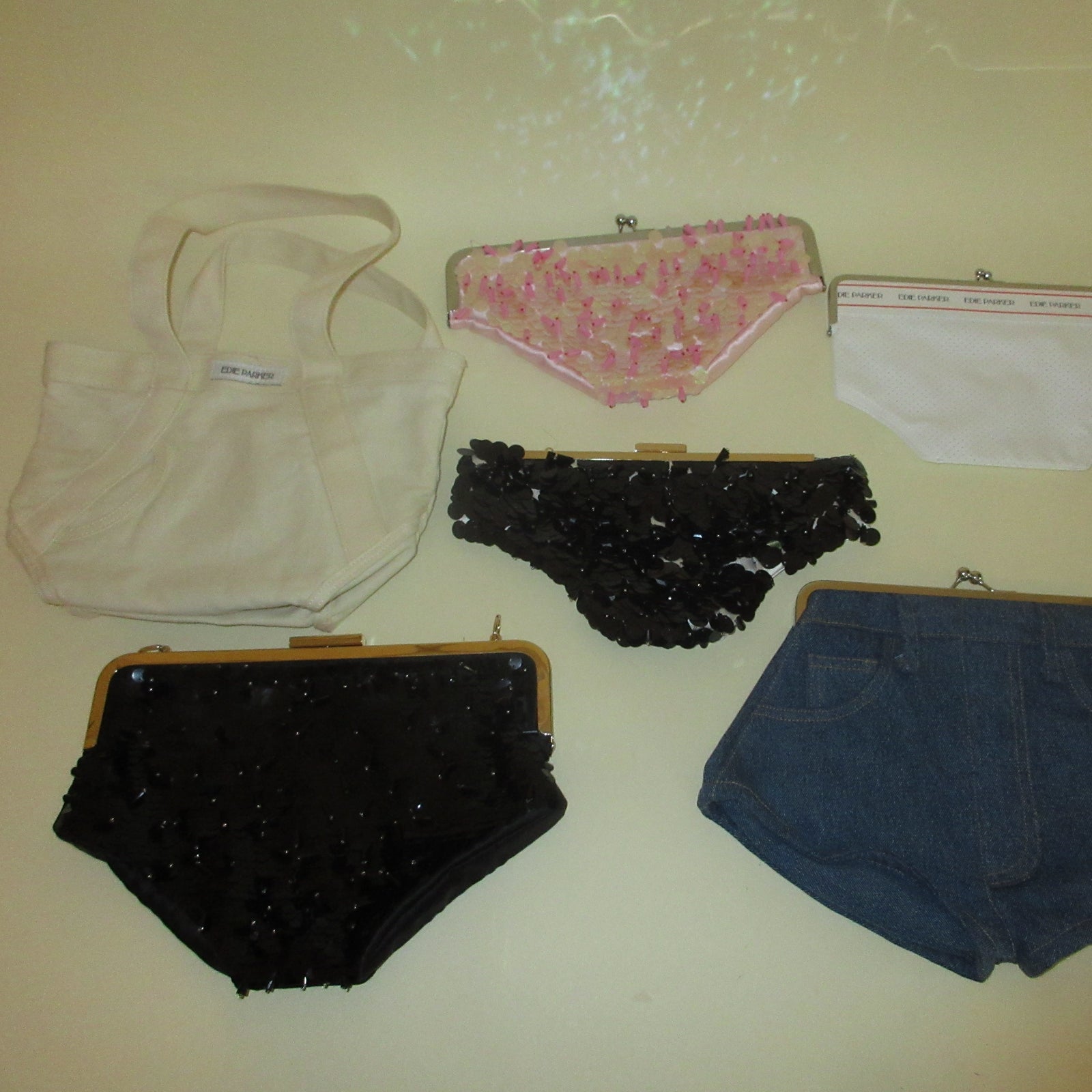 A collection of six women's underwear and shorts items arranged on a beige background, including a cream canvas tote bag, a pink floral bikini-style bottom, a plain white bottom, a black ruffled bottom, a black sequined bottom, and blue denim shorts.