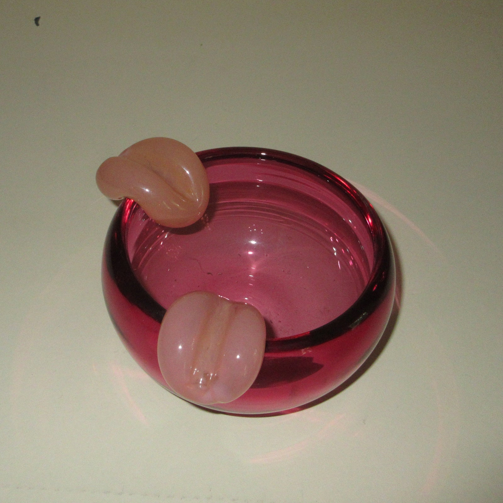 A small pink glass bowl with three protruding pink glass loops, photographed on a plain off-white surface.